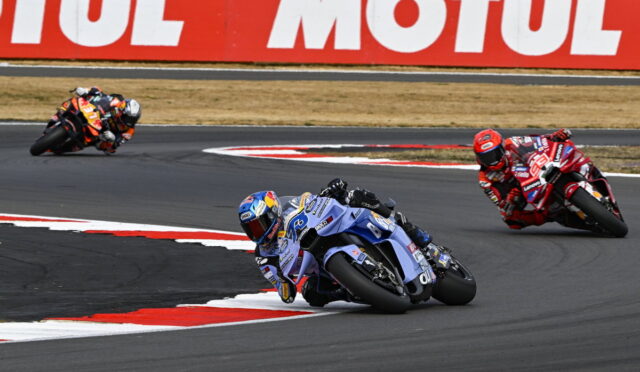 Motorcycle race taking a sharp left turn; blue bike leads, red bikes behind on a curving track with a large MOTUL banner in the background.