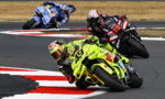 Leading rider in neon-yellow motorcycle leans into a left-handed turn on a racetrack, with two followers behind him on a curved course.
