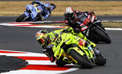 Leading rider in neon-yellow motorcycle leans into a left-handed turn on a racetrack, with two followers behind him on a curved course.