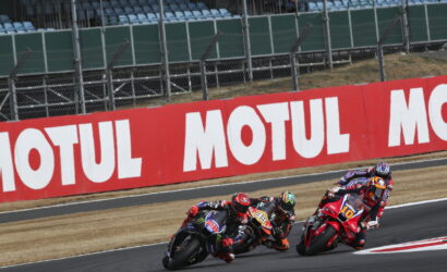 Motorcycle racers lean into a corner on a race track with a red MOTUL barrier in the background, three bikes in a tight pack