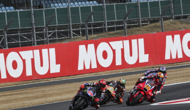 Motorcycle racers lean into a corner on a race track with a red MOTUL barrier in the background, three bikes in a tight pack