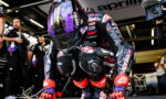 MotoGP rider in full racing suit crouches in the pit lane, helmeted and ready, with crew and a camera operator in the background.