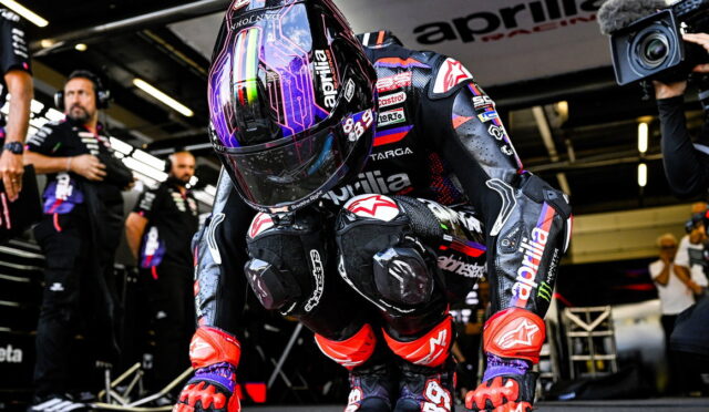 MotoGP rider in full racing suit crouches in the pit lane, helmeted and ready, with crew and a camera operator in the background.