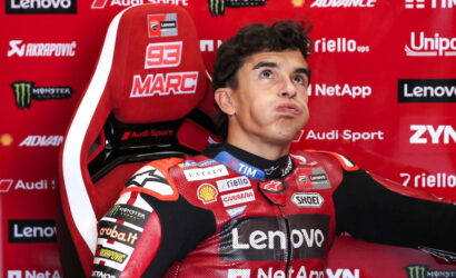 MotoGP rider in a red Lenovo racing suit sits in a sponsor-filled red backdrop, puffing his cheeks playfully in a press photo.