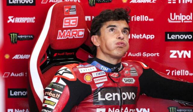 MotoGP rider in a red Lenovo racing suit sits in a sponsor-filled red backdrop, puffing his cheeks playfully in a press photo.