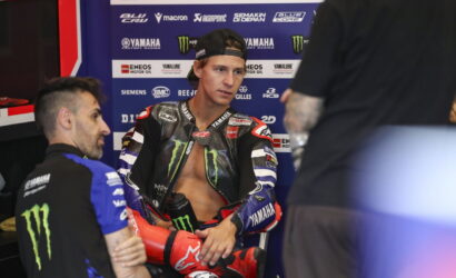 MotoGP rider in Yamaha gear sits in a pit area, chatting with a team member against a sponsor backdrop with Monster Energy logos.
