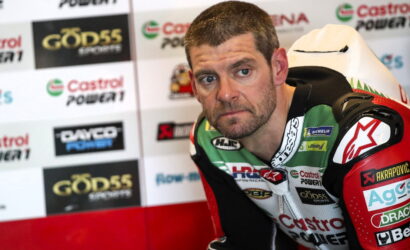 Motorcycle racer in green, white, and red suit sits in a paddock with sponsor banners in the background, looking toward the camera with a focused expression
