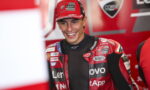 MotoGP rider in a red Lenovo Ducati racing suit smiling in the paddock/garage area.