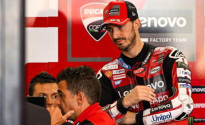MotoGP rider in red and black Lenovo racing suit with sponsor patches stands in a team garage, talking with two engineers nearby in a red paddock backdrop.