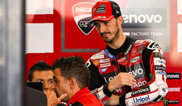 MotoGP rider in red and black Lenovo racing suit with sponsor patches stands in a team garage, talking with two engineers nearby in a red paddock backdrop.