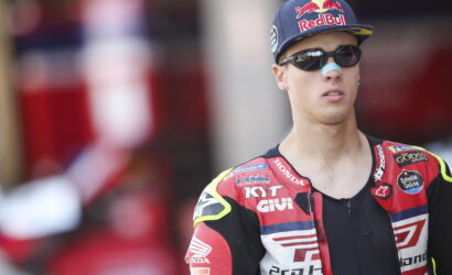 Portrait of a male motorcycle racer wearing a Red Bull cap, sunglasses, and a red Honda racing suit with sponsor logos on a blurred background.