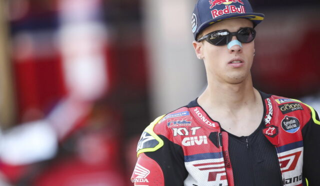 Portrait of a male motorcycle racer wearing a Red Bull cap, sunglasses, and a red Honda racing suit with sponsor logos on a blurred background.