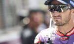 Moto racer wearing sunglasses and cap, drinking from a bottle with a straw during a break at the track, purple suit visible near the chest area