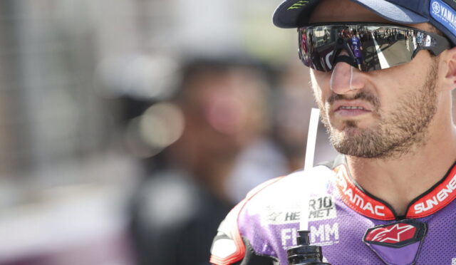 Moto racer wearing sunglasses and cap, drinking from a bottle with a straw during a break at the track, purple suit visible near the chest area