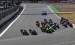 MotoGP race on a curved track with riders leaning into a turn; pit wall on the left and a police car with blue lights in the background.