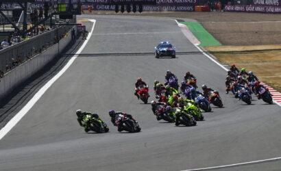 MotoGP race on a curved track with riders leaning into a turn; pit wall on the left and a police car with blue lights in the background.