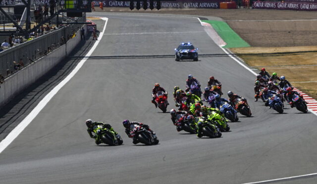 MotoGP race on a curved track with riders leaning into a turn; pit wall on the left and a police car with blue lights in the background.