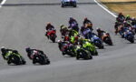 Group of motorcycle racers leaning into a turn on a race track, tightly packed and speeding through the corner.