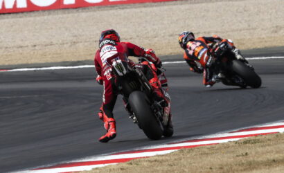Two motorcycle racers lean into a sharp curve on a race track, foreground rider in red leading the orange-clad rider in the background, both bikes cornering aggressively.