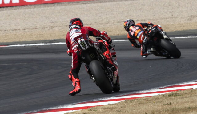 Two motorcycle racers lean into a sharp curve on a race track, foreground rider in red leading the orange-clad rider in the background, both bikes cornering aggressively.