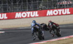 Two motorcycle racers lean into a sharp turn on a racetrack, blue and black bikes close together near a Motul advertisement barrier.