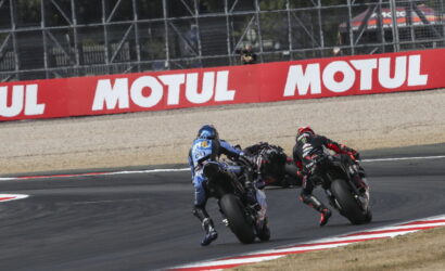 Two motorcycle racers lean into a sharp turn on a racetrack, blue and black bikes close together near a Motul advertisement barrier.