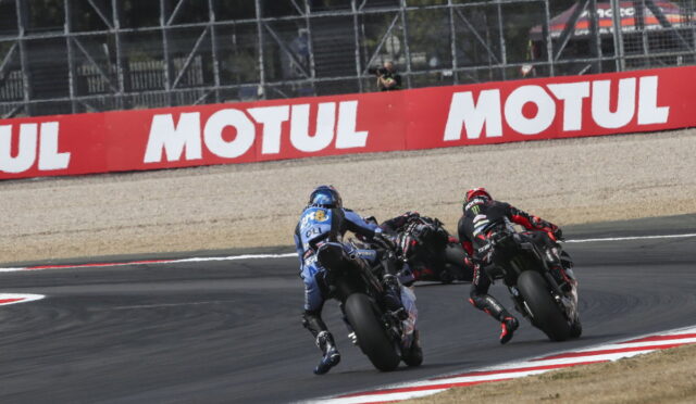 Two motorcycle racers lean into a sharp turn on a racetrack, blue and black bikes close together near a Motul advertisement barrier.