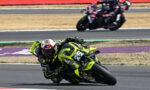 Motorcycle racer in neon yellow bike leaning into a turn on a racetrack, number 23, with a blurred second rider in the background.