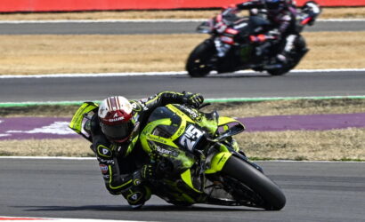 Motorcycle racer in neon yellow bike leaning into a turn on a racetrack, number 23, with a blurred second rider in the background.