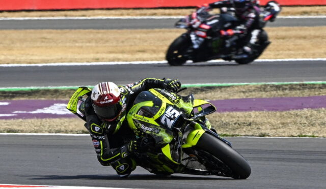 Motorcycle racer in neon yellow bike leaning into a turn on a racetrack, number 23, with a blurred second rider in the background.