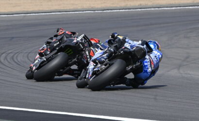 Three sport motorcycles lean into a left-hand turn on a race track, riders tucked in racing posture with rear tires smoking slightly for grip (blue, black, and red bikes).
