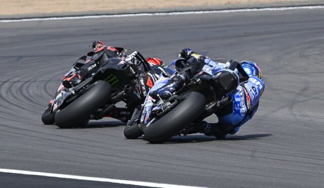 Three sport motorcycles lean into a left-hand turn on a race track, riders tucked in racing posture with rear tires smoking slightly for grip (blue, black, and red bikes).