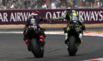 Two sport motorcycle riders on a race track touch gloves while leaning into a left-hand turn, spectators behind the barrier.
