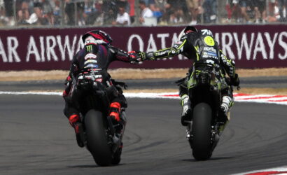 Two sport motorcycle riders on a race track touch gloves while leaning into a left-hand turn, spectators behind the barrier.