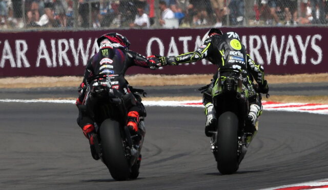 Two sport motorcycle riders on a race track touch gloves while leaning into a left-hand turn, spectators behind the barrier.