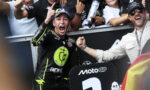 MotoGP rider in black neon-yellow gear cheers with fist raised as teammate grips his arm in celebration, MotoGP sign visible in foreground.