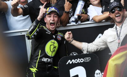 MotoGP rider in black neon-yellow gear cheers with fist raised as teammate grips his arm in celebration, MotoGP sign visible in foreground.