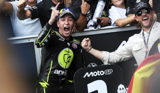 MotoGP rider in black neon-yellow gear cheers with fist raised as teammate grips his arm in celebration, MotoGP sign visible in foreground.