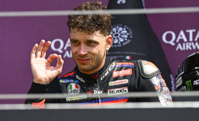 MotoGP rider in a black-and-red leather suit on a podium, making an OK gesture with his right hand.