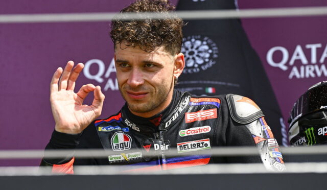 MotoGP rider in a black-and-red leather suit on a podium, making an OK gesture with his right hand.