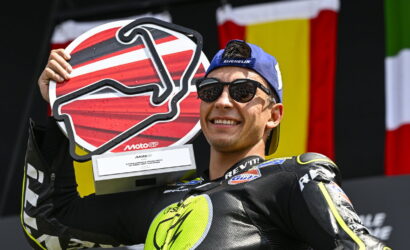 Rider in a black neon racing suit wearing a blue cap and sunglasses, proudly holding a large red-and-white trophy on stage with flags in the background.