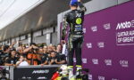 MotoGP rider in a black and yellow racing suit stands on his bike at the podium as fans cheer and photograph him in front of a purple Qatar Airways backdrop.