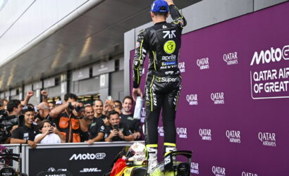 MotoGP rider in a black and yellow racing suit stands on his bike at the podium as fans cheer and photograph him in front of a purple Qatar Airways backdrop.
