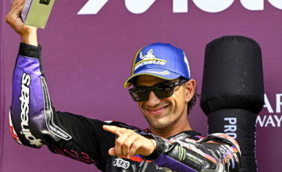 Male motorcycle racer in a blue Michelin cap and sunglasses raises a trophy on a purple podium backdrop, smiling at the camera.