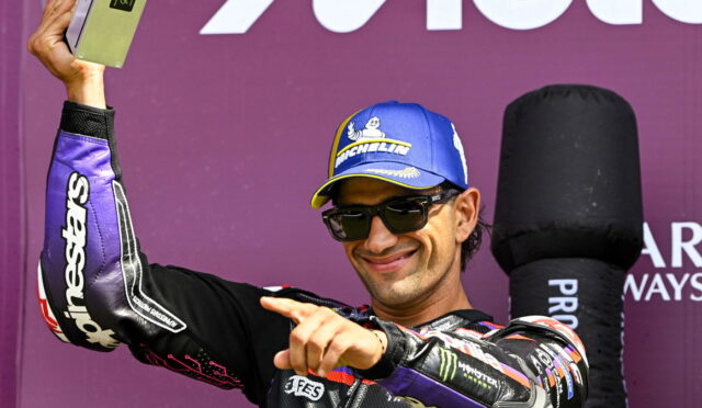 Male motorcycle racer in a blue Michelin cap and sunglasses raises a trophy on a purple podium backdrop, smiling at the camera.