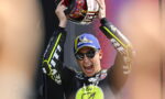 Motorcycle racer on a podium raising a helmet in celebration, wearing a blue Michelin cap and sunglasses.