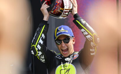 Motorcycle racer on a podium raising a helmet in celebration, wearing a blue Michelin cap and sunglasses.