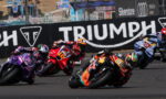 Group of motorcycle racers leaning into a sharp turn on a race track with a large Triumph barrier in the background.