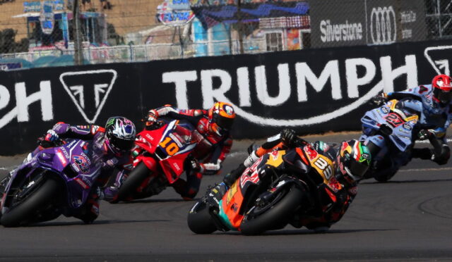 Group of motorcycle racers leaning into a sharp turn on a race track with a large Triumph barrier in the background.