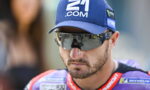 Male motorcycle racer wearing a blue cap with 21.COM and reflective sunglasses, focused on the race ahead.
